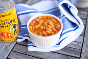 Mustard-Q Baked beans can next to beans in white dish on a table with a blue and white towel